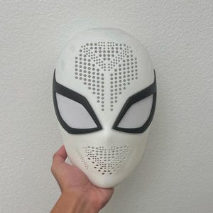 May include: A white full-face mask featuring a perforated pattern across the forehead and chin area. The mask includes two large, angular eye openings outlined in black, with white mesh lenses covering the eye sockets for visibility.