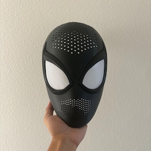 May include: A matte black full-face mask featuring large, almond-shaped white mesh eye lenses. The mask has a perforated texture across the forehead and mouth area, with a smooth, contoured surface design that covers the entire face.