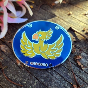 May include: A blue and yellow enamel pin featuring a cartoon Chocobo, a bird-like creature from the Final Fantasy video game series. The pin has a silver border and the word "Chocobo" written in silver lettering.