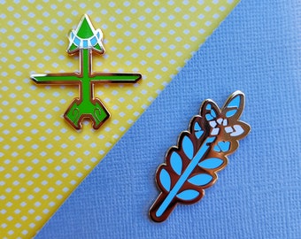 May include: Two enamel pins with gold-colored borders. One pin is green with a geometric design, and the other is a blue and white leaf design. The pins are on a yellow polka dot and blue background.