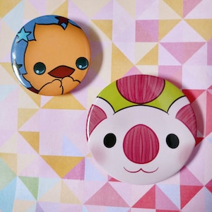 May include: Two round pins with cartoon animal faces. The first pin is blue, yellow, and white with a bird-like creature with a star on its head. The second pin is green, pink, and white with a pig-like creature with a pink nose.