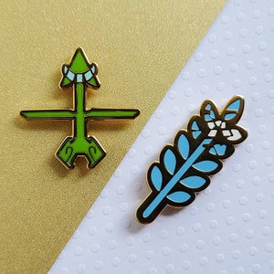 May include: Two enamel pins with gold trim. One is a green arrow design, the other is a blue and white leaf. The pins are on a gold and white background.