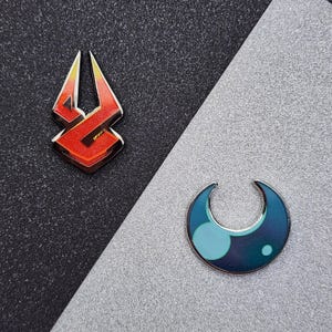 Puede incluir: Dos pines esmaltados. Un pin es un diseño estilizado rojo y naranja con un contorno plateado. El otro pin tiene forma de media luna azul con un círculo y un punto azul claro. Los pines están sobre un fondo negro y plateado.