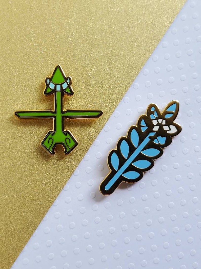 May include: Two enamel pins with gold trim. One is a green arrow design, the other is a blue and white leaf. The pins are on a gold and white background.