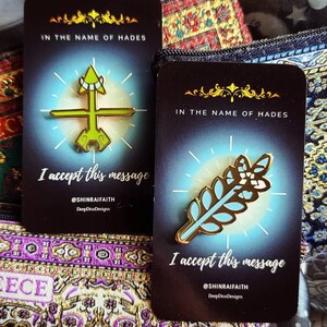 May include: Two enamel pins on cards with the text "IN THE NAME OF HADES" and "I accept this message." One pin is a green and gold arrow design, the other is a gold and light blue leaf design. The background is a mix of patterned fabrics.