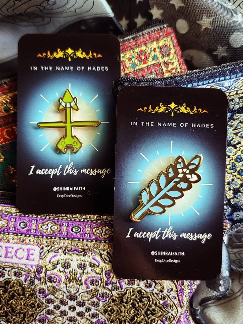 May include: Two enamel pins on cards with the text "IN THE NAME OF HADES" and "I accept this message." One pin is a green and gold arrow design, the other is a gold and light blue leaf design. The background is a mix of patterned fabrics.