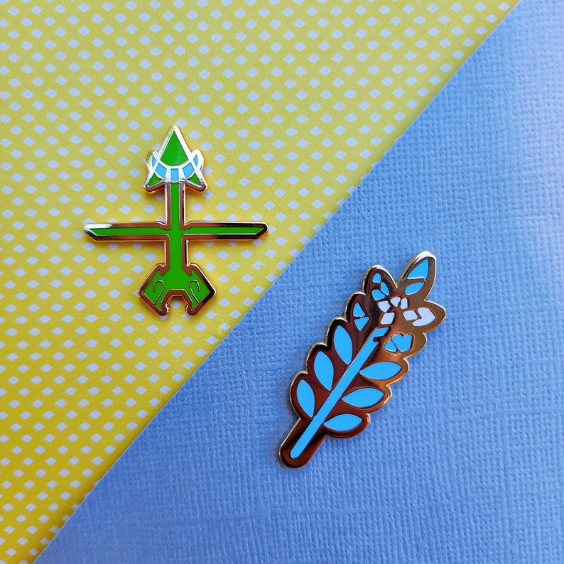 May include: Two enamel pins with gold-colored borders. One pin is green with a geometric design, and the other is a blue and white leaf design. The pins are on a yellow polka dot and blue background.