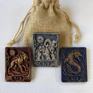 May include: Three rectangular tokens with raised relief designs and a burlap drawstring pouch. The left token is red with a creature, the center is silver with a mammoth, and the right is blue with a sea serpent. Each token features the text 3/3.