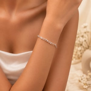 May include: A delicate silver-toned bracelet featuring three thin, layered chains adorned with small, evenly spaced metallic beads. The bracelet wraps around the wrist, showcasing a refined, multi-strand design with a polished finish and a secure clasp.