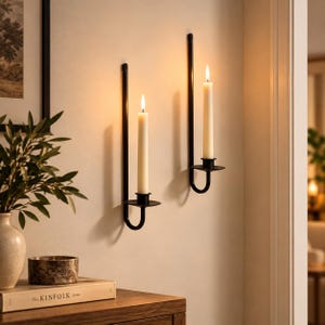 May include: Two black metal wall-mounted candle holders with long, slender vertical arms and curved bases. Each holder supports a single off-white taper candle with a lit flame, casting a warm glow against the light-coloured wall surface.