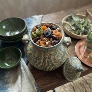 Puede incluir: Un tarro de cerámica con dos asas y tapa a juego, decorado con intrincados patrones verdes y beige, lleno de una mezcla de albaricoques secos, dátiles, higos, nueces y pistachos, acompañado de tres cuencos verdes y una cesta tejida.