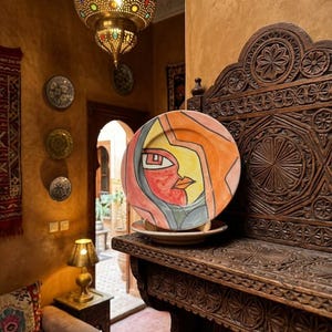 Può includere: Un piatto rotondo in ceramica con un'illustrazione stilizzata e astratta di un volto nei toni dell'arancione, giallo, rosso e grigio. Il piatto è esposto su un piccolo supporto sopra una mensola in legno scuro finemente intagliata.