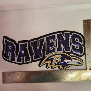 May include: A sequined Ravens patch featuring purple and black lettering with a gold, purple, and white raven logo. A metal ruler placed along the bottom and right side shows the patch is approximately 9.5 inches wide and 4 inches tall.