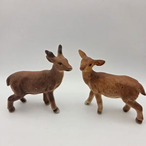 May include: Two small deer figurines crafted from brown ceramic. The left deer features short, dark-tipped antlers, while the right deer has smooth, pointed ears. Both figures are depicted in a standing position with realistic shading and textures.