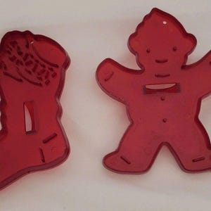 May include: Two red plastic cookie cutters. One cutter is shaped like a Christmas stocking with a textured cuff and a rectangular cutout. The other cutter is shaped like a gingerbread person with a rounded head, outstretched arms, and a rectangular cutout.