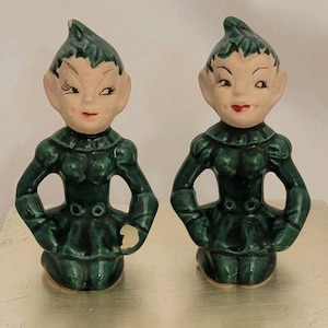 May include: Two ceramic elf figurines with pointed ears and hats, wearing dark green outfits with ruffled collars and waist details. The figures have pale skin, painted facial features, and one figure shows a small chip on the left hand.