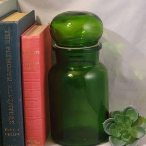 May include: A cylindrical green glass apothecary jar with a rounded, matching green glass lid. The jar is positioned next to a blue book titled The Plymouth Adventure, a red book titled The Arts, and a small green artificial succulent plant.