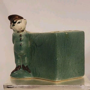 Peut inclure: Un pot de fleurs en céramique représentant un petit elfe portant un chapeau et des chaussures marron. L'elfe est vêtu d'une combinaison verte avec trois boutons foncés sur le devant. Le récipient rectangulaire attaché a une finition verte mouchetée.
