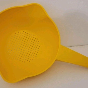 Puede incluir: Un colador de plástico de color amarillo brillante que presenta un cuenco redondo con una base perforada para drenar líquidos. El colador incluye un mango largo y plano con un pequeño orificio circular en el extremo y un pico vertedor.