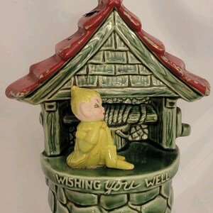 May include: A ceramic wishing well figurine featuring a red roof with chipped paint, a green stone-textured base, and a yellow elf figure sitting in front. The front of the well is embossed with the text WISHING YOU WELL in raised lettering.