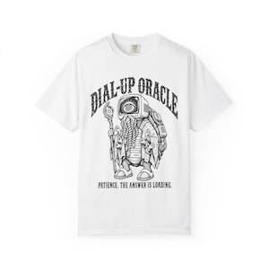 Puede incluir: Una camiseta blanca con un gráfico negro de una criatura con cabeza de monitor de ordenador que muestra un ojo, múltiples brazos y un caparazón. El texto dice DIAL-UP ORACLE arriba y PATIENCE. THE ANSWER IS LOADING. debajo de la ilustración.