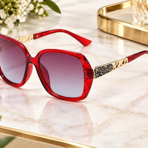 May include: Oversized sunglasses with a translucent red plastic frame featuring a faceted edge design. The temples are gold-toned metal with a cutout geometric pattern and a section of black crystals near the hinges. The lenses have a dark gradient.