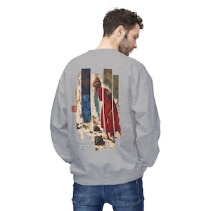 May include: A heather grey crewneck sweatshirt featuring a graphic print on the back. The design displays a figure in a red robe standing within vertical panels, accompanied by blue geometric patterns and the text ENKIDU PREMIERE.