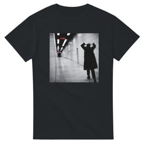 Może przedstawiać: Czarny t-shirt z krótkim rękawem z kwadratowym nadrukiem graficznym na klatce piersiowej. Nadruk przedstawia czarno-białe zdjęcie osoby w długim płaszczu idącej przez wyłożony kafelkami tunel z oświetleniem sufitowym i małym czerwonym tekstem.