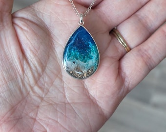 Op de afbeelding: Een druppelvormige hanger met een harsinleg die een oceaanlandschap nabootst. Het ontwerp verloopt van diepblauw aan de bovenkant naar turkoois en wit schuim, eindigend met een laagje zand aan de onderkant, gevat in een zilveren frame.