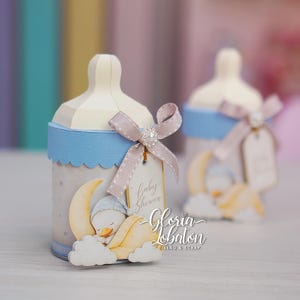 May include: A decorative baby bottle favor featuring a light blue scalloped band, a beige ribbon bow with a central faux pearl, and a tag reading "baby shower". The bottle displays a graphic of a duckling sleeping on a crescent moon and clouds.
