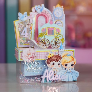 May include: A multi-layered decorative box featuring a Cinderella theme with a light blue carriage, a silver glitter slipper, a clock tower, and two cartoon characters. The front displays the name Alicia and the number 4 with pink and blue accents.