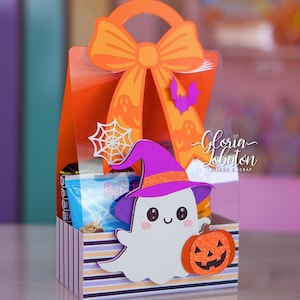 以下が含まれることがあります： ハロウィーンをテーマにしたギフトボックス。透明なプラスチック製の本体に、リボンの形をしたオレンジ色の紙製ハンドルが付いています。紫色の帽子をかぶった白い幽霊、キラキラしたオレンジ色のカボチャ、銀色のクモの巣、小さな紫色のコウモリで装飾されています。