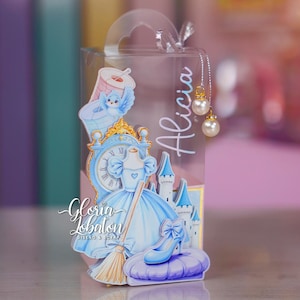 Puede incluir: Una caja de regalo de plástico transparente con un recorte decorativo de un vestido de gala azul, un reloj, un castillo, un pájaro y un zapato de cristal. El nombre Alicia está impreso en el lateral y dos dijes de perlas cuelgan del asa.