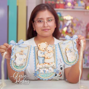 Puede incluir: Un estandarte decorativo con tres paneles festoneados en tonos azul claro y crema. Los paneles muestran ilustraciones de un body de bebé, una familia de tres ositos de peluche y un biberón. El texto "IT'S A BOY!" aparece en el panel central.