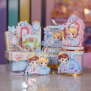 Puede incluir: Una colección de cajas decorativas para fiestas con ilustraciones de un príncipe y una princesa, una carroza de calabaza y una torre de reloj. Los artículos tienen tonos azul pastel y rosa, detalles dorados brillantes y el nombre Alicia.