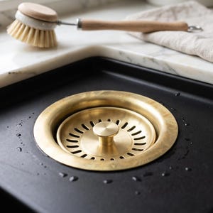 Può includere: Un filtro circolare in ottone per scarico di lavello da cucina con pomello centrale e bordo fessurato, inserito in una vasca nera opaca. Lo scarico presenta una finitura oro metallizzato ed è circondato da piccole gocce d'acqua.