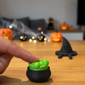 May include: A small black cauldron with a bright green bubbling liquid insert. Beside it sits a black witch hat with a simple face design. The cauldron is round with a smooth finish, and the green insert features a textured, bubbly surface.