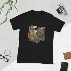 May include: A black t-shirt featuring a graphic illustration of two green aliens in a living room. One alien sits on a sofa watching a television labelled Earth Channel, while the other stands nearby. Speech bubbles contain the text: watching? and Earth Channel.