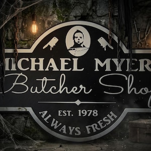 Può includere: Un cartello rettangolare nero con bordi arrotondati che presenta il testo bianco MICHAEL MYERS Butcher Shop. Sopra il testo c'è un grafico circolare di un volto e due mannaie. Sotto il testo c'è la frase EST. 1978 ALWAYS FRESH.