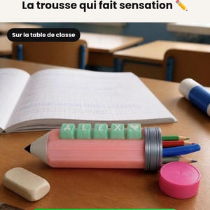 Op de afbeelding: Een roze etui in de vorm van een potlood met een grijze dop en een rij lichtgroene vierkante toetsen die ALEX spellen met een hartsymbool. Het etui bevat verschillende kleurpotloden en wordt vergezeld door een witte rechthoekige gum.
