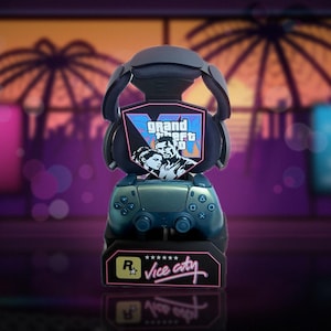May include: A dark blue gaming controller stand featuring a Grand Theft Auto logo graphic with a character illustration. The base displays the Rockstar Games logo and the text Vice City in a stylized pink font, holding a metallic blue controller.