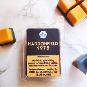 Könnte beinhalten: Ein schwarzer Behälter für Wachs-Schmelzen mit einem weißen Etikett, auf dem "The Salty Hive Handmade Candles" und "Haddonfield 1978" in fetter Schrift stehen. Das Etikett enthält auch den Text "artisan wax melts", "crisp fall air, apple bobbing, pumpkin carving & trick or treating. death has come to your little town", "thesaltyhiveandbody.com", "net wt. 3 oz | 85 g", "SMALL BATCH, HANDCRAFTED IN AKRON, OHIO".