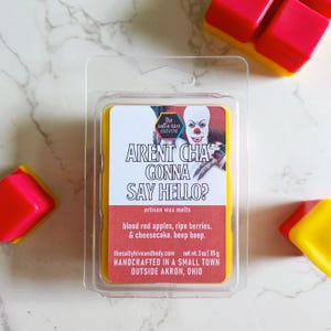 Puede incluir: Un paquete de plástico transparente que contiene una cera perfumada amarilla y roja. La cera perfumada tiene un gráfico de un payaso y el texto "ARENT CHA GONNA SAY HELLO?". El paquete también incluye el texto "artisan wax melts" y "blood red apples, ripe berries, & cheesecake. beep beep."