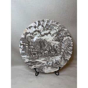 May include: A round ceramic plate with a scalloped edge featuring a black and white transferware scene of a horse-drawn carriage in front of a timber-framed building. The detailed illustration includes trees, a small stream, and figures.