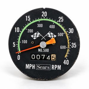 May include: A round black analog gauge featuring a dual-scale display for MPH and RPM. The face includes white numbers, a green and yellow arc, two crossed checkered flags, the text NO.500, Sears, and a mechanical odometer reading 00745.