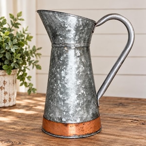 Könnte beinhalten: Ein verzinkter Metallkrug mit konischem Körper, breitem Ausguss und geschwungenem Griff. Der Hauptkörper hat eine gesprenkelte graue Oberfläche, während die Basis mit einem glatten, polierten kupferfarbenen Band am Boden akzentuiert ist.