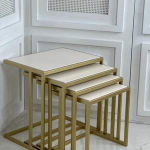 Pode incluir: Um conjunto de quatro mesas de encaixe com tampos retangulares em branco sujo e estruturas com acabamento em ouro metálico. Cada mesa apresenta um design geométrico limpo com quatro pernas verticais ligadas por uma base.