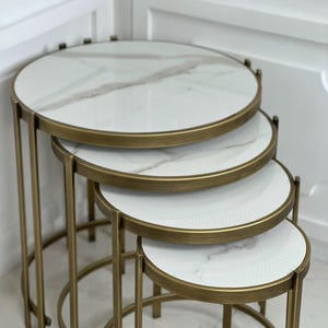 May include: A set of four nesting tables with circular white marble-patterned tops and brushed brass-toned metal frames. Each table features a thin, circular rim and slender vertical legs that curve into a base, arranged in descending size order.