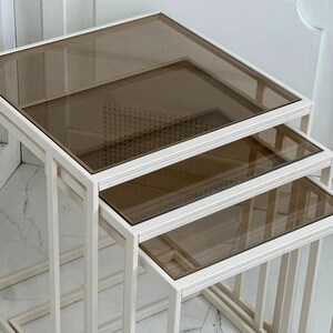 Peut inclure: Un ensemble de trois tables gigognes dotées de cadres rectangulaires avec une finition blanc cassé mat. Chaque table est surmontée d'un panneau en verre brun translucide et lisse. Les tables sont conçues pour s'emboîter les unes dans les autres.