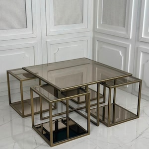 Puede incluir: Un conjunto de mesas nido que incluye una mesa central cuadrada y tres mesas cuadradas más pequeñas. Cada mesa tiene un marco metálico en tono bronce y una superficie de cristal tintado marrón, creando un conjunto geométrico.